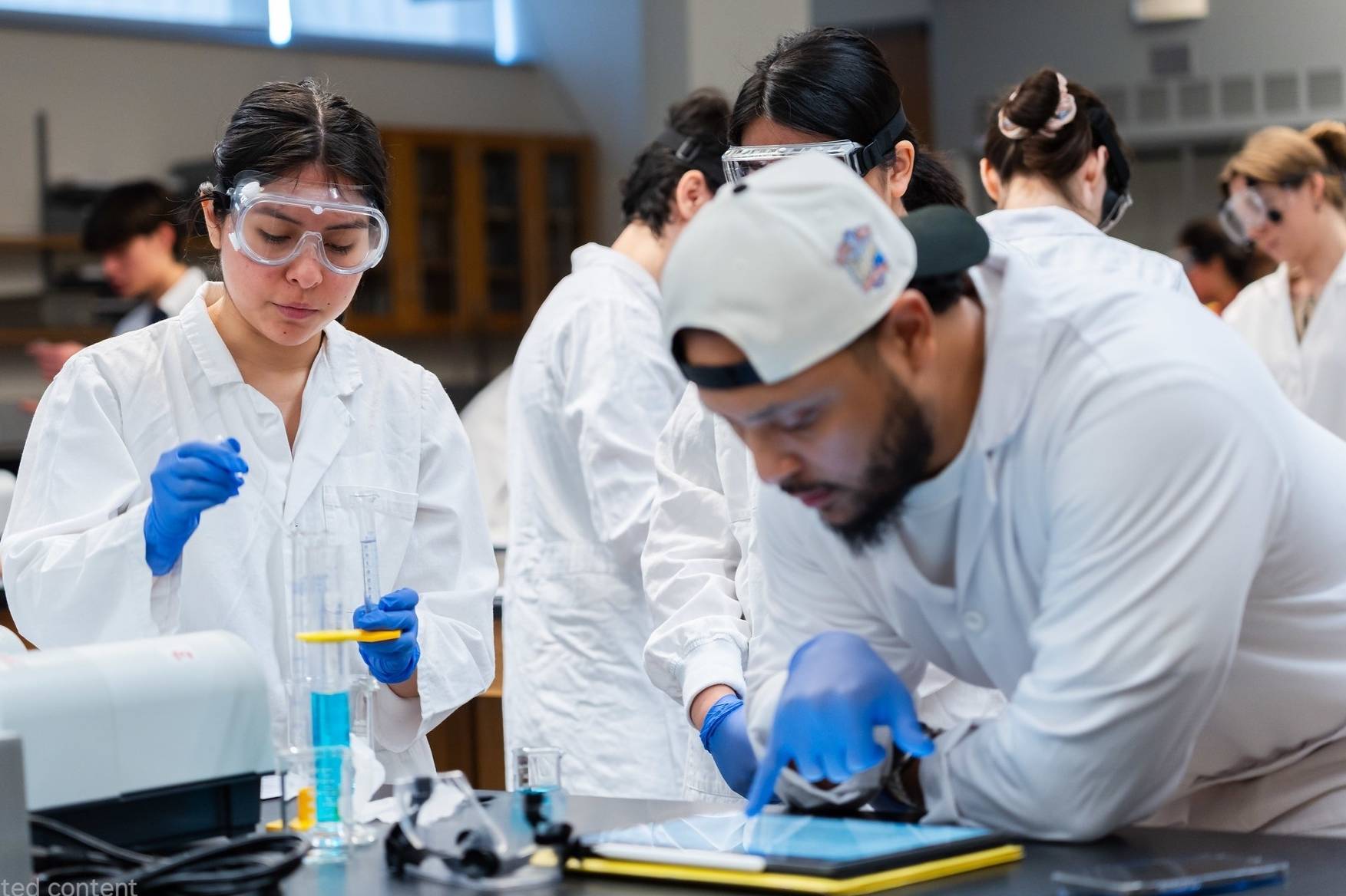San Diego Mesa College students in a science lab wearing goggles and gloves conduct an experiment while one student references a tablet.